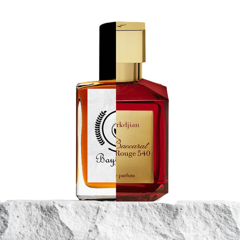 The image displays a split composition of two perfume bottles merged together. The left side represents Baytulkenz Perfumes, with its signature amber-colored liquid and a white label featuring the brand's logo. The right side shows the iconic red bottle of Baccarat Rouge 540 by Maison Francis Kurkdjian, with its deep red hue and gold label. Both bottles are placed on a stone-textured surface, creating a visually engaging contrast between the two luxury perfumes (BR540)